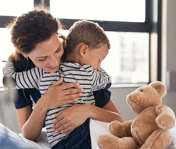 A woman embraces a young child in a comforting hug while a teddy bear sits nearby.